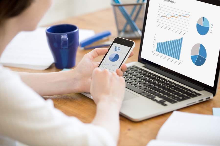 Businesswoman using smartphone and laptop with charts and graphs on the screen.