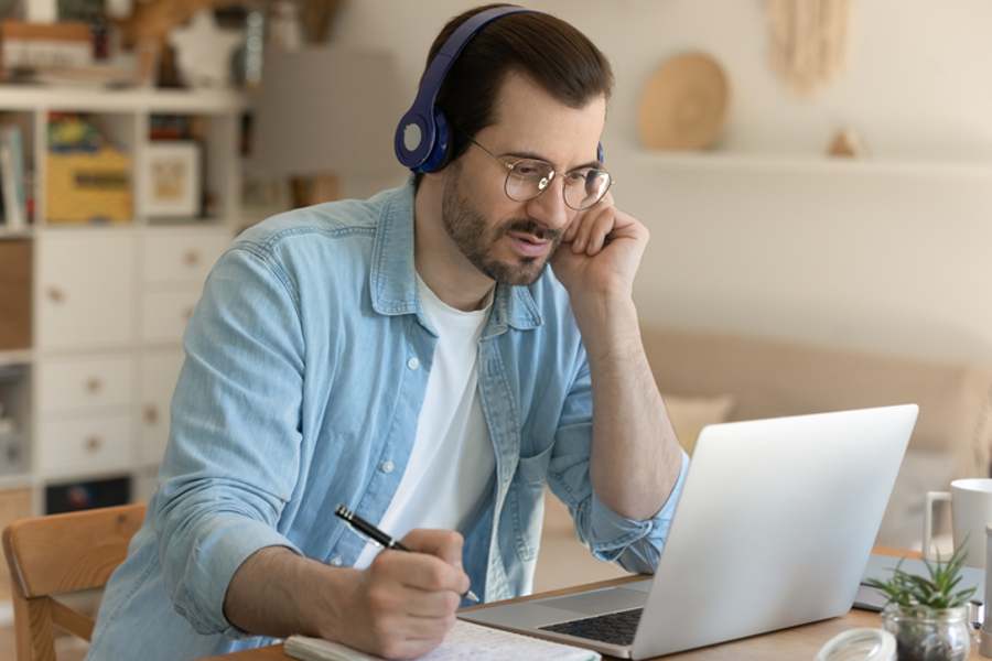 男人戴着耳机坐在笔记本电脑旁听着，做着笔记