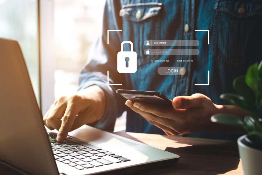 Man's hand enters his login information on a laptop while holding his phone in the other hand.